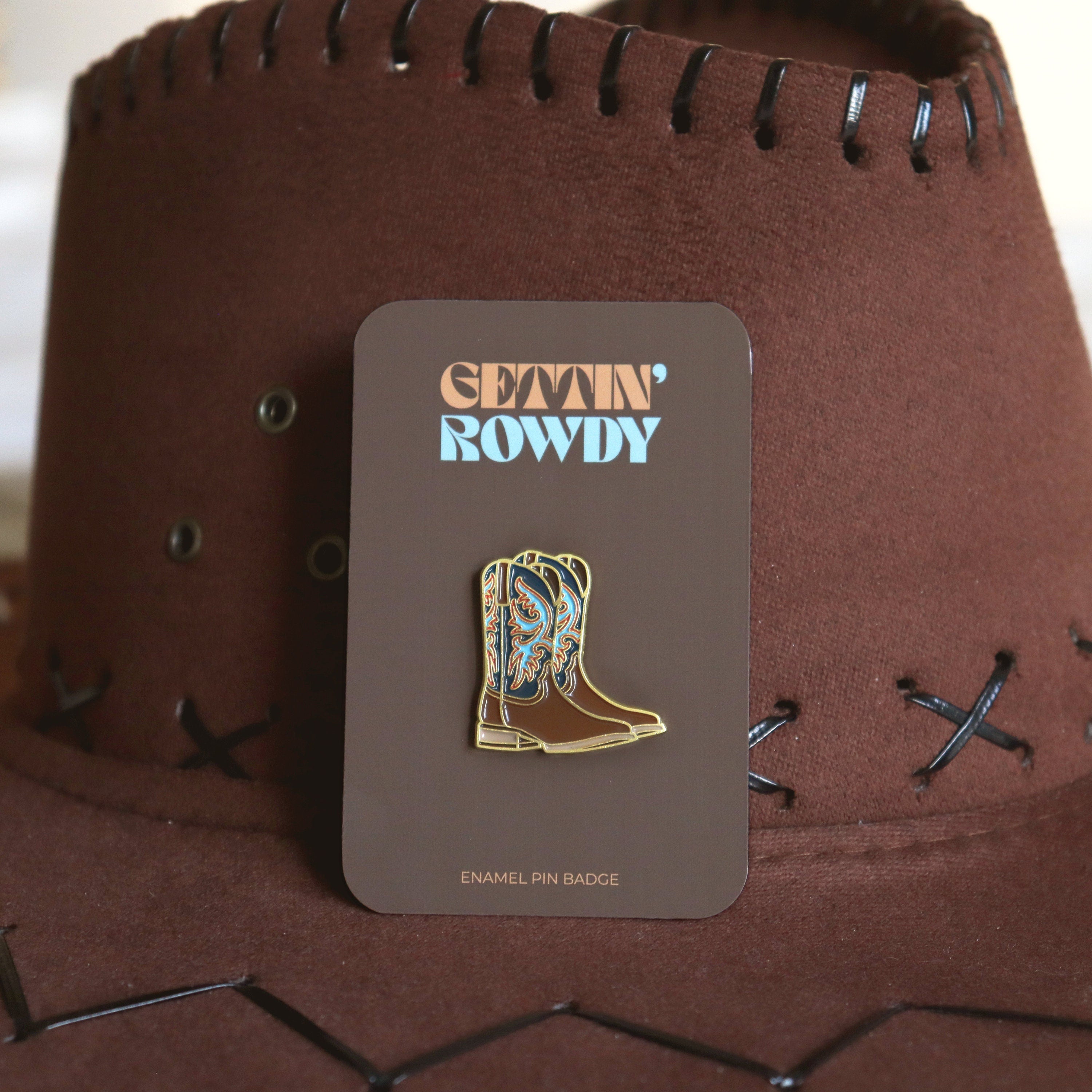 Cowboy boot enamel pin badge on a brown card with 'Gettin' Rowdy' text, against a brown cowboy hat