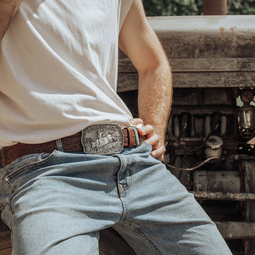 A close-up lifestyle shot of a man wearing a brown leather belt with a large, detailed silver-toned buckle featuring a cowboy riding a bucking horse. The belt is worn with a brown patterned leather strap over light-wash blue jeans and a white t-shirt, set against a rustic, weathered metal background.