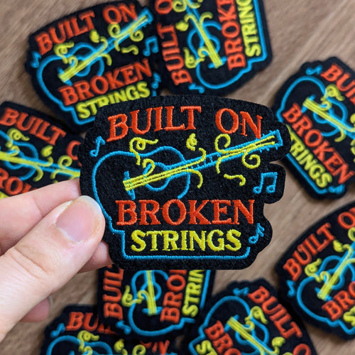 Colorful 'Built on Broken Strings' patches with a hand holding one patch.