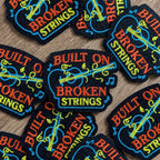 Embroidered patches with 'Built on Broken Strings' text and guitar design on a wooden surface.