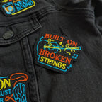 Black jacket with colorful patches including 'Built on Broken Strings' and 'Neon Bound'.