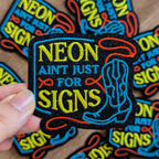Colorful patch with 'NEON AIN'T JUST FOR SIGNS' text held by a hand, surrounded by similar patches on a wooden surface.