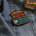 Two embroidered patches on a denim background, one reading 'Whiskey Bent and Neon Bound' and the other 'Built on Broken Strings'.